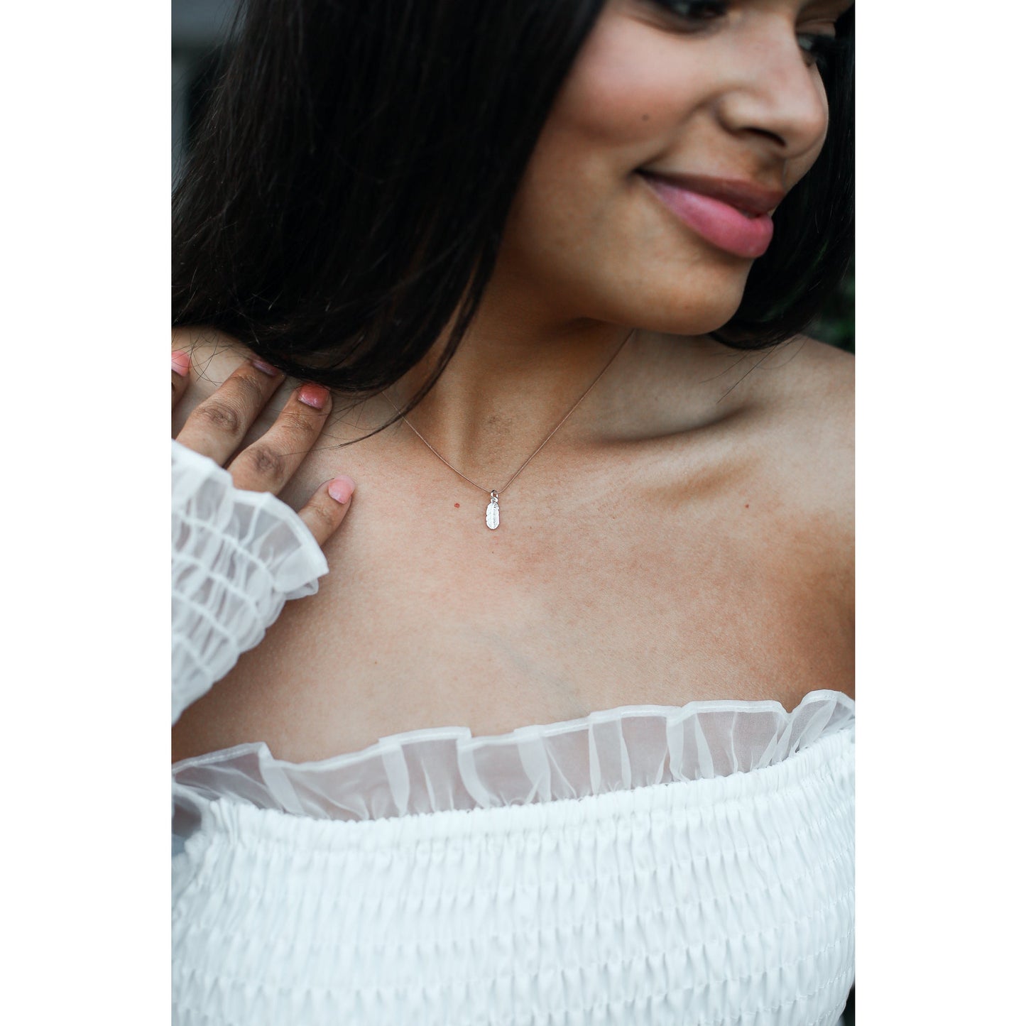 A woman wearing an off-the-shoulder white top and a delicate necklace smiles softly.