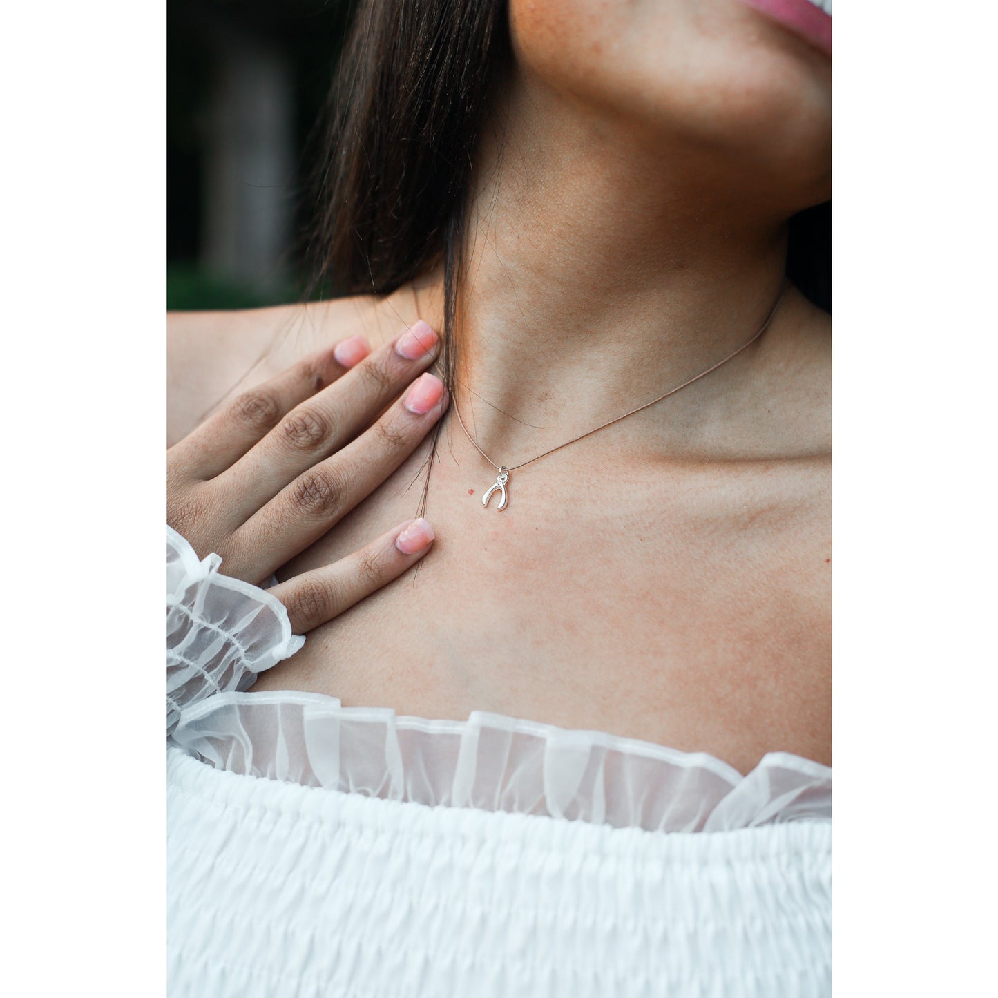 A woman wearing a white top and a wishbone necklace touches her chest.