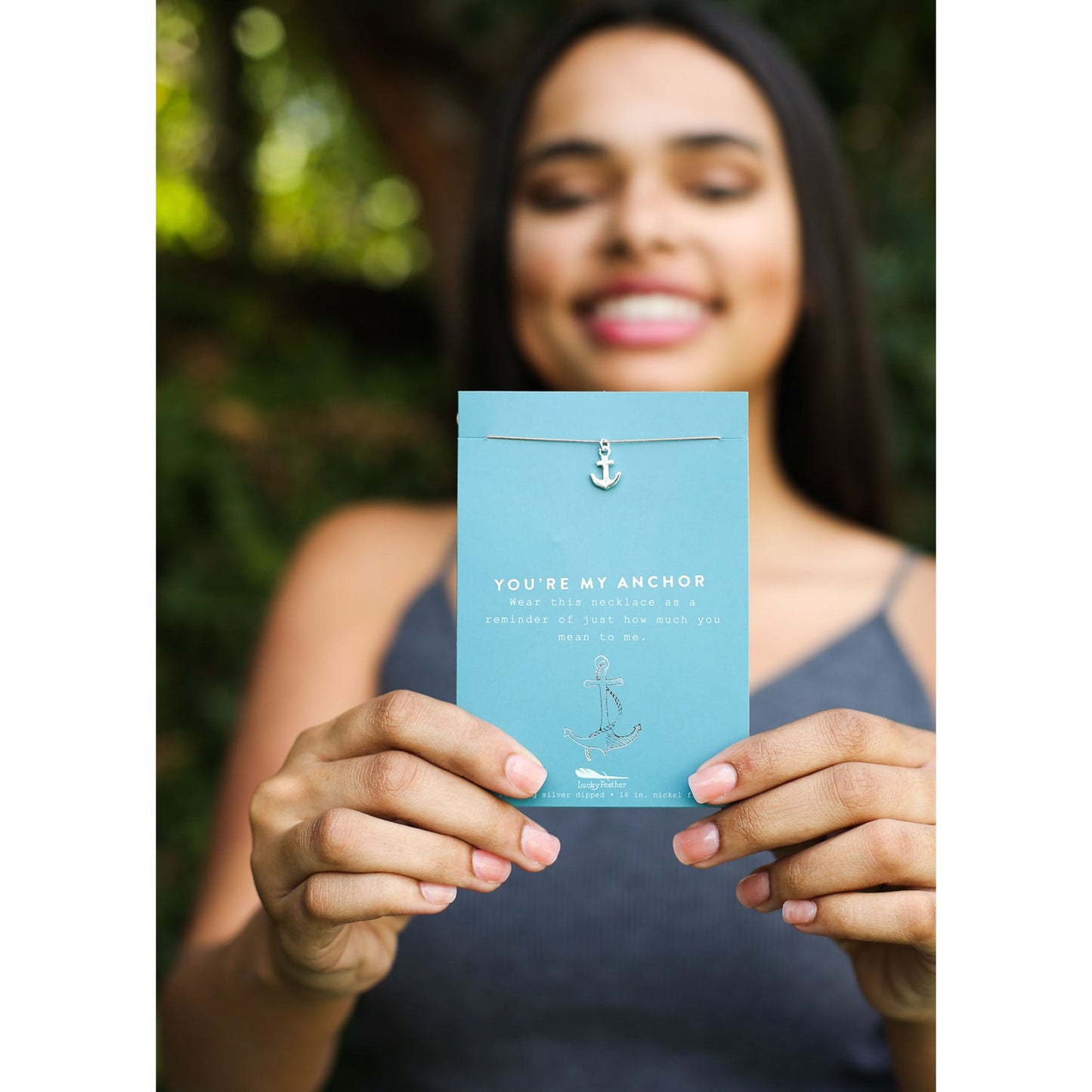 A blurry woman smiles while holding a light blue card with an anchor necklace.